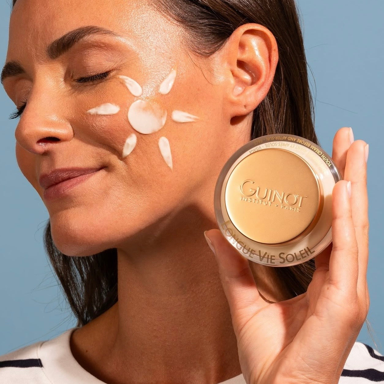 Longue Vie Soleil Visage Guinot, pot tenu dans la main d'une femme. Dessin d'un soleil fait sur le visage de la femme à l'aide de la crème.